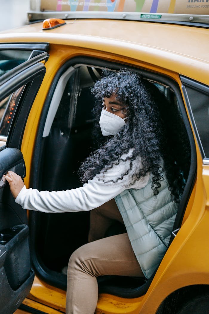 Side view emotionless African American female in warm clothes and face mask opening door and getting off yellow taxi